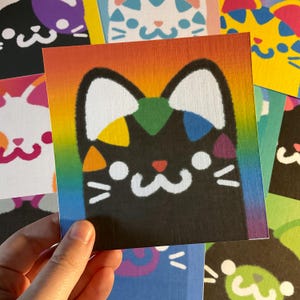 May include: A square art print featuring a black cat face with white ears, cheeks, and mouth. The cat has colourful geometric shapes in green, blue, purple, orange, and yellow on its head. The background is a vibrant vertical rainbow gradient.