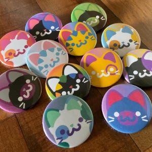 May include: A selection of cat-themed button badges in various colours. Each badge displays a different cat face design, with rainbow, blue, yellow, and purple colour schemes. The badges are round and have a glossy finish.