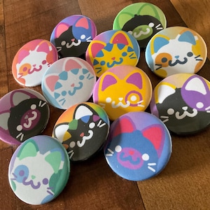 May include: A selection of round cat-themed badges in various colours. Each badge displays a different cat design, with colour schemes including pink, blue, yellow, and rainbow patterns. The badges are arranged on a wooden surface.