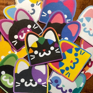 May include: A selection of cat-shaped stickers in various colours. The stickers feature different colour combinations, including black, yellow, blue, and purple. White accents are used for the eyes and whiskers. The stickers are arranged on a brown surface.