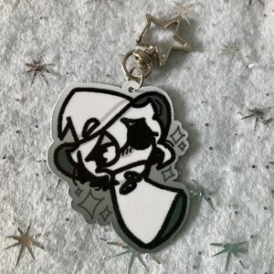 May include: A clear acrylic keychain featuring a black and white cartoon character with a hat and bow tie. The keychain has a silver star-shaped clasp and a silver ring. The character is outlined in black and has a white background.