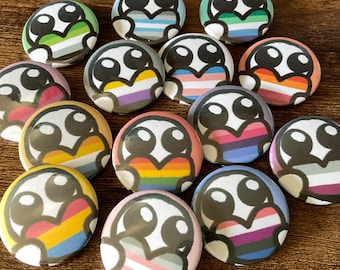 May include: A collection of circular pin-back buttons featuring a cartoon character with large black eyes holding a heart. Each heart displays a different multi-colored striped pattern representing various pride flags in vibrant shades of red, orange, yellow, green, blue, purple, pink, and white.