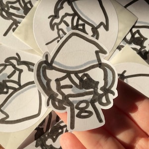 May include: A hand holding a white sticker with a black outline drawing of a person wearing a hat. The person's face is visible, and the number "18" is below the face. Other similar stickers are in the background.
