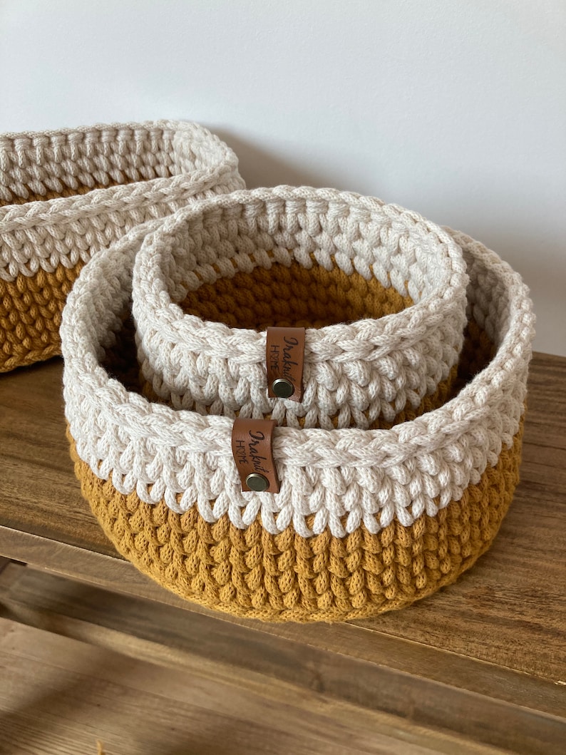 K&ouml;nnte beinhalten: Zwei geh&auml;kelte K&ouml;rbe, einer im anderen, sind aus wei&szlig;em und senfgelbem Garn gefertigt. Beide K&ouml;rbe haben ein braunes Leder-Etikett mit der Aufschrift "Draped Home".