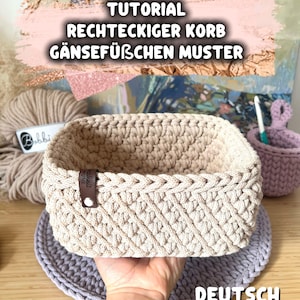 Puede incluir: Una cesta rectangular beige de ganchillo con un patrón texturizado. La cesta tiene una etiqueta de cuero y se muestra sobre una alfombra de ganchillo de color púrpura claro. La imagen incluye el texto "TUTORIAL RECHTECKIGER KORB GÄNSEFÜßCHEN MUSTER" y "DEUTSCH".