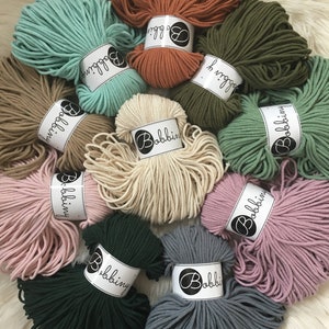 Op de afbeelding: Assortiment van 10 spoelen katoenen touw in verschillende kleuren, waaronder groen, blauw, roze, beige en oranje. Elke spoel is gelabeld met de merknaam "Bobbiny".