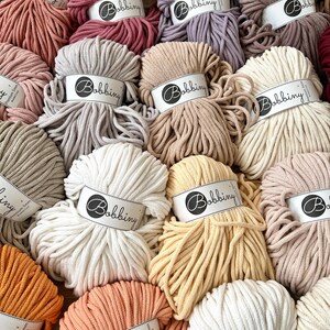 Op de afbeelding: Een selectie van katoenen touw in verschillende kleuren, elk met een label met een witte cirkel met daarin het woord "Bobbiny". Het touw is in een cirkelvormige opstelling gerangschikt.