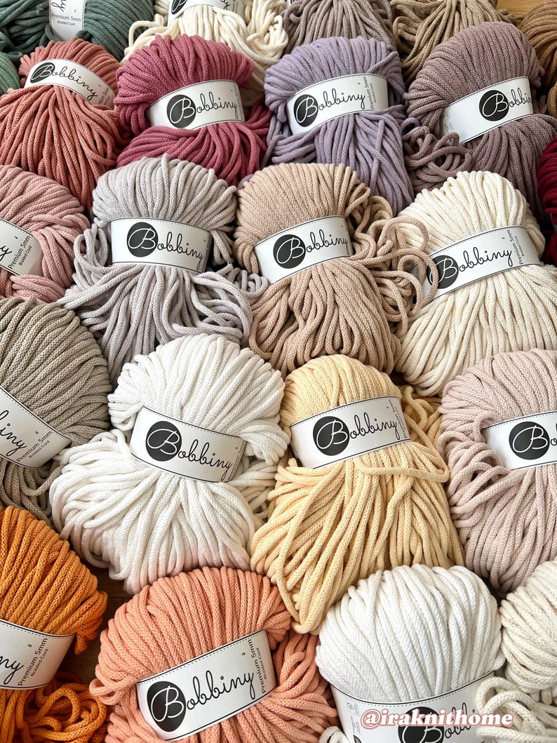 Op de afbeelding: Een selectie van katoenen touw in verschillende kleuren, elk met een label met een witte cirkel met daarin het woord "Bobbiny". Het touw is in een cirkelvormige opstelling gerangschikt.