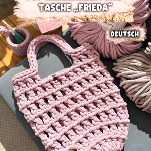 Könnte beinhalten: Eine handgefertigte, rosa gehäkelte Tragetasche mit einem gewebten Design. Die Tasche hat einen geflochtenen Griff und einen Holzknopfverschluss. Das Bild zeigt auch Spulen aus rosa und beigem Garn, einen kleinen gehäkelten Korb und den Text "PDF HÄKELANLEITUNG TASCHE FRIEDA" und "DEUTSCH".