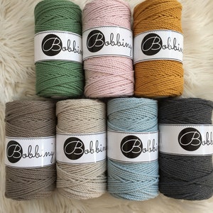 Può includere: Otto bobine di corda di cotone in vari colori, tra cui verde, rosa, giallo senape, beige, azzurro e grigio. Ogni bobina è etichettata con la parola "Bobbiny".