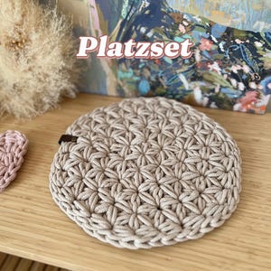 Op de afbeelding: Een ronde, beige gehaakte placemat met een stervormig patroon. De placemat ligt op een houten oppervlak. Het woord "Platzset" is zichtbaar op de achtergrond.