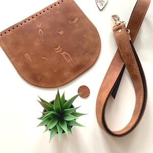 Könnte beinhalten: Eine braune Ledertasche mit Katzenmotiv und einem passenden braunen Lederriemen. Die Tasche ist unfertig und hat Löcher am Rand zum Nähen.
