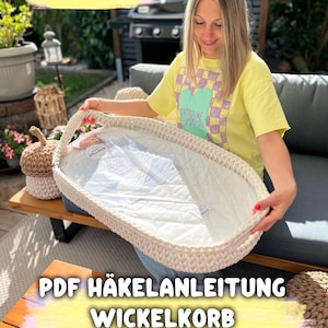 Könnte beinhalten: Ein cremefarbener, ovaler Babykorb aus Häkelarbeit mit Griff. Der Korb enthält einen weißen, gepolsterten Einsatz. Der Text "PDF HÄKELANLEITUNG WICKELKORB" ist in Gelb und Weiß dargestellt. Weitere Häkelartikel sind ebenfalls zu sehen.