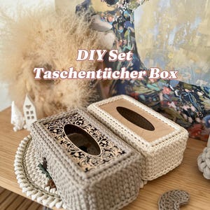 Könnte beinhalten: Zwei gehäkelte Taschentuchbox-Bezüge, einer beige und einer weiß, mit einem Holzsockel. Der beige Bezug hat ein dekoratives Blumenmuster. Der Text "DIY Set Taschentücher Box" ist im Bild sichtbar.