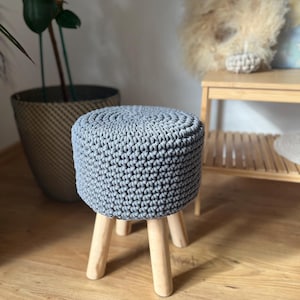 Op de afbeelding: Een grijze gehaakte kruk met houten poten. De kruk staat op een houten vloer.