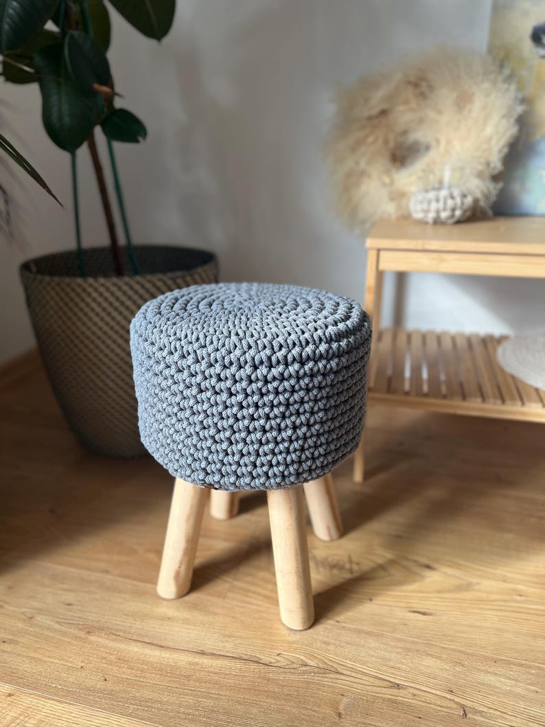 Op de afbeelding: Een grijze gehaakte kruk met houten poten. De kruk staat op een houten vloer.