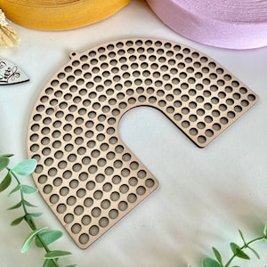 Könnte beinhalten: Ein halbkreisförmiges Bastelstück aus hellem Holz mit vielen kleinen, gleichmäßig verteilten Löchern. Das Stück hat eine abgerundete Form und einen flachen Boden. Im Hintergrund sind Rollen mit gelbem, hellblauem und lila Band zu sehen.