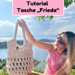 Könnte beinhalten: Eine cremefarbene, handgehäkelte Tasche mit offenem Knotenmuster und zwei integrierten Tragegriffen. Ein rosafarbenes rechteckiges Schild über der Tasche zeigt den schwarzen Text: Tutorial Tasche „Frieda“. Die Tasche hat ein kleines dunkles Etikett.