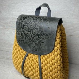 May include: A yellow crocheted backpack with a black leather flap featuring an embossed turtle design. The backpack has a top handle and adjustable straps.