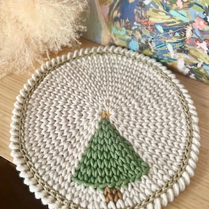 Puede incluir: Un posavasos redondo de punto color crema con un diseño de árbol de Navidad verde. El árbol tiene un tronco marrón y una estrella amarilla. El posavasos está bordeado con una cuerda dorada y mide aproximadamente 15 cm de diámetro.