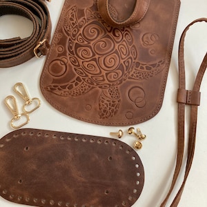 Könnte beinhalten: Eine braune Ledertasche mit einem Schildkröten-Design, einem langen braunen Lederriemen, einem kleineren braunen Lederstück mit Löchern zum Nähen und goldenen Metallbeschlägen.