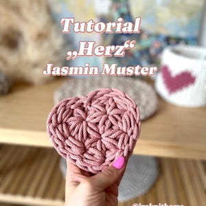May include: A pink heart-shaped crocheted coaster with a floral pattern. The coaster is being held by a hand. The text "Tutorial "Herz" Jasmin Muster" is visible above the coaster.