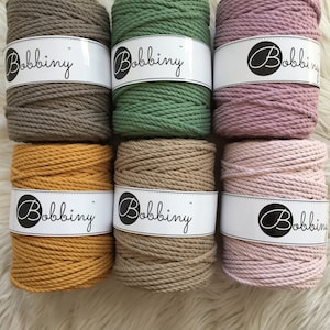 Può includere: Sei rotoli di corda di cotone in vari colori, tra cui marrone, verde, rosa, giallo, beige e rosa chiaro. Ogni rotolo ha un'etichetta bianca con la parola "Bobbiny" stampata su di essa.