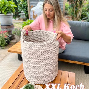 Pode incluir: Uma grande cesta bege tricotada à mão com duas alças. A cesta é cilíndrica e está sobre uma mesa de madeira. Uma pequena cesta de malha combinando com uma planta está em um sousplat redondo bege. O texto "XXL Korb" é visível.