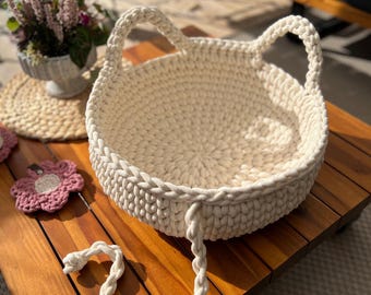 May include: A white crocheted cat bed with two handles. The bed is sitting on a wooden table with a pink crocheted flower on the table. The text "@iraknithome Katzen Korb" is visible in the image.