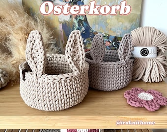 May include: Two beige crocheted baskets with bunny ear handles. The baskets are on a wooden shelf with a pink crocheted flower and a pink crocheted heart on the lower shelf. The text "Tutorial Osterkorb" is visible above the baskets.