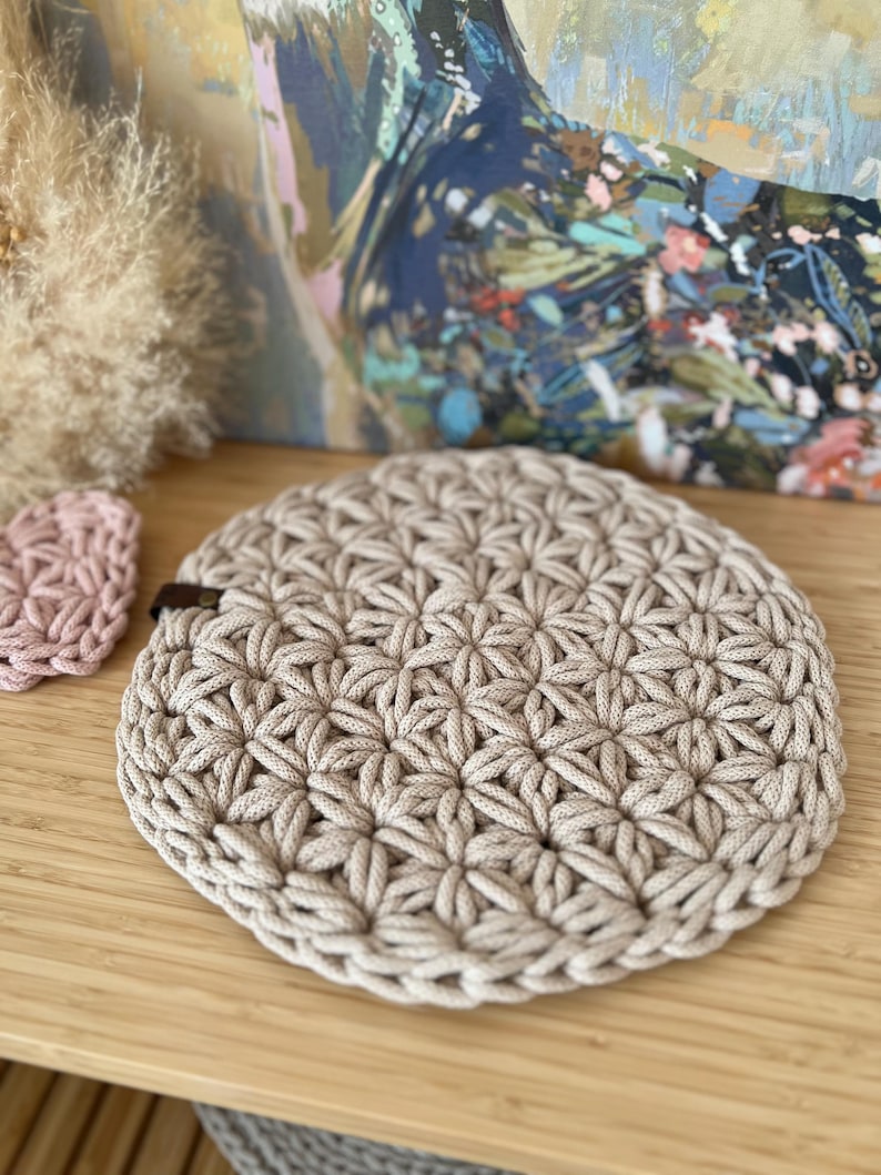 Op de afbeelding: Een ronde, beige gehaakte onderzetter met een stervormig patroon. De onderzetter is gemaakt van dik katoenen touw en heeft een bruine leren lus.