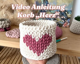 Puede incluir: Una cesta de crochet blanca con un diseño de corazón rosa. La cesta está etiquetada "Video Anleitung Korb,Herz".