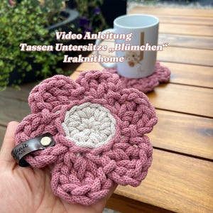 May include: A pink, flower-shaped coaster with a beige centre, made from thick, braided yarn. A small leather tag is attached. The text "Video Anleitung Tassen Untersetze Blümchen Iraknithome" is visible in the background. The coaster is designed to hold a mug.