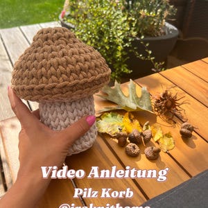 May include: A crocheted mushroom-shaped basket with a brown top and a light beige bottom. The basket is sitting on a wooden table with acorns and leaves scattered around it. The text "Video Anleitung Pilz Korb @iraknithome" is written on the table.