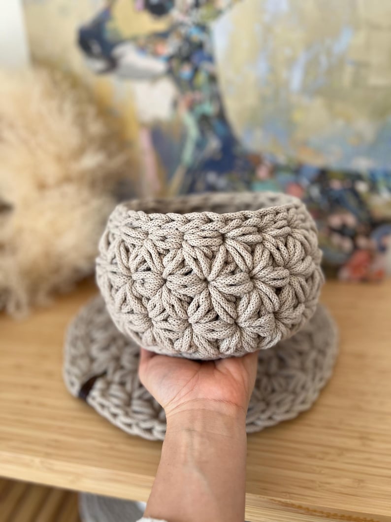 Op de afbeelding: Een beige gehaakte mand met een stervormig patroon. De mand staat op een beige gehaakte mat.