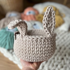 Op de afbeelding: Een beige gehaakte mand met twee konijnenoren. De mand is gemaakt van dik, getextureerd garen en heeft een ronde vorm.