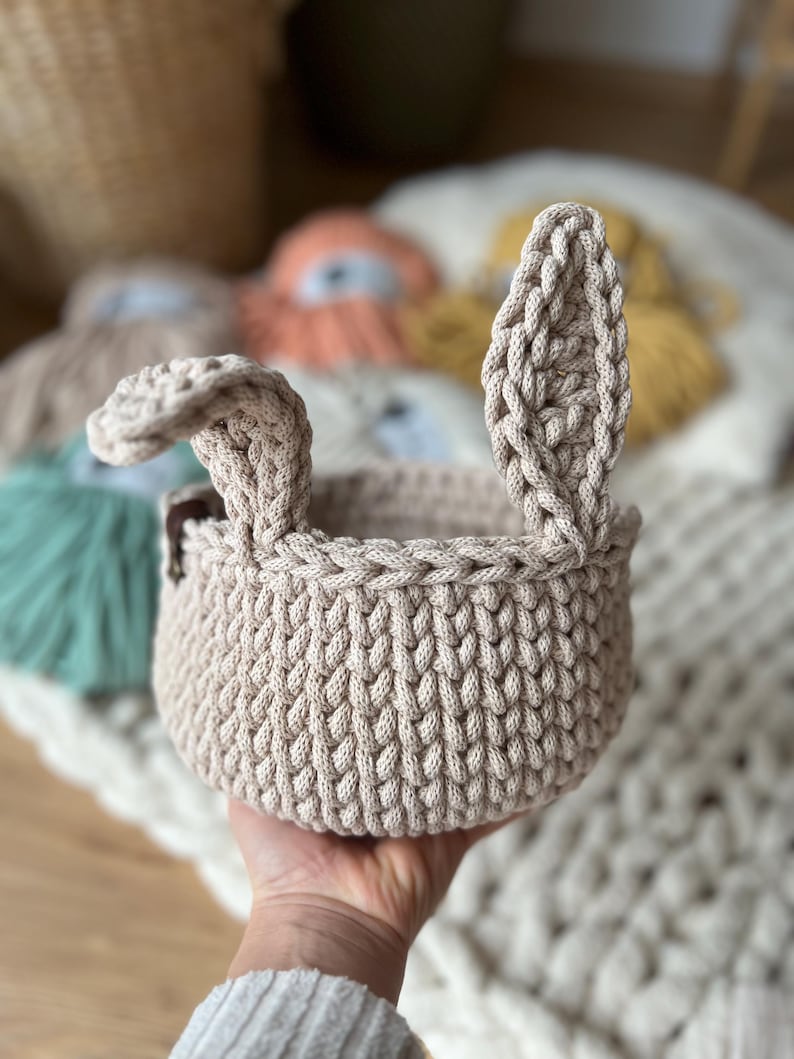 Op de afbeelding: Een beige gehaakte mand met twee konijnenoren. De mand is gemaakt van dik, getextureerd garen en heeft een ronde vorm.