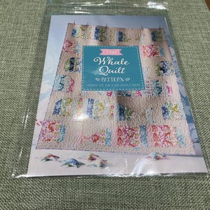 May include: A Tilda Whale Quilt pattern booklet in a clear plastic sleeve. The cover features a light-colored quilt with whale-shaped blocks in various floral patterns, a blue central label with white text, and small whale-shaped fabric cutouts.