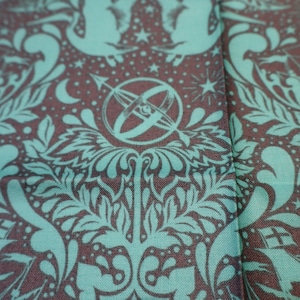 May include: A woven fabric featuring a repeating pattern in teal and dark brown. The design includes illustrations of sailing ships, celestial stars, crescent moons, and a central armillary sphere motif surrounded by stylized leafy foliage.
