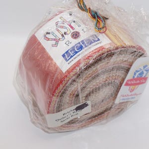 May include: A roll of fabric, wrapped in clear plastic, featuring a variety of colors including red, brown, and beige. The packaging includes labels with text and a decorative string.