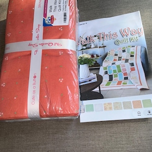 May include: A wrapped fabric bundle with a floral pattern in shades of orange and white, labeled "Fat Quarter Shop" and "Cute as a Button." Accompanied by a "Walk This Way Quilt Kit" booklet featuring a colorful quilt design.