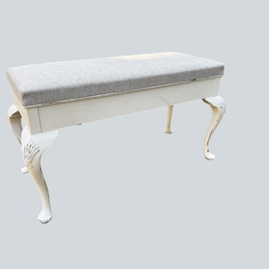 May include: A rectangular bench featuring a light grey upholstered seat with a textured fabric finish. The base is finished in a glossy white paint and is supported by four curved legs that feature carved shell-like details at the top.