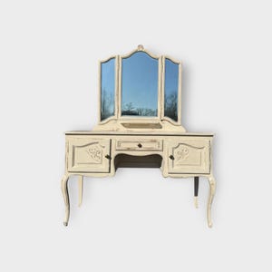 May include: Cream-coloured vanity unit with a tri-fold mirror. The vanity has two cabinets, a drawer, and a central opening. The legs are curved, and the mirror reflects a blue sky and trees. It has decorative carvings.