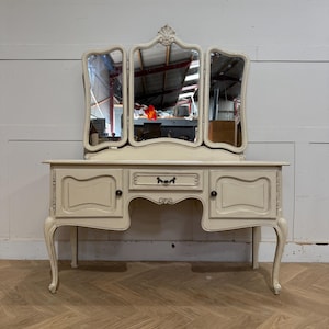 May include: Cream-coloured vanity unit with a tri-fold mirror. The vanity has a drawer and two cabinet doors, with ornate detailing and curved legs. The mirrors have a decorative top. The unit is set against a light-coloured wall.