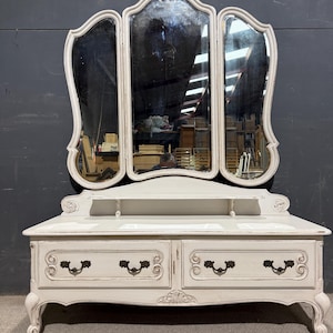 May include: A white vanity table with a three-panel mirror. The table has two drawers with ornate handles and carved details. The mirror has a scalloped edge and is framed in white.