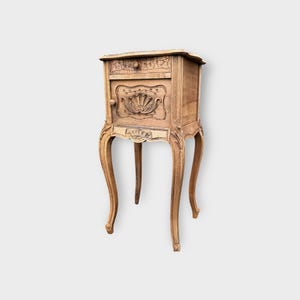 May include: An antique light brown bedside cabinet with a drawer and a small door. The cabinet has decorative carvings, including a shell design on the door and floral patterns on the drawer. The curved legs add to its vintage appeal.