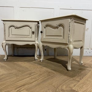 May include: A pair of off-white bedside cabinets with decorative features. Each cabinet has a single door with a detailed panel and a dark knob. The cabinets have curved legs and are on a wooden floor.