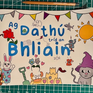 Könnte beinhalten: Eine bunte Illustration eines Buches mit dem Text "Ag Dathlú tríd an Bhliain" auf weißem Hintergrund. Die Illustration zeigt einen Schneemann, eine Rose, ein Kleeblatt, eine Sonne, einen Roboter, eine Katze mit einem lila Hut, eine Sandburg, eine Krabbe und einen Drachen.