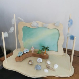 May include: A wooden beach playset featuring a painted backdrop of a sky and sea. Includes toy cars, ice cream, a tent, a palm tree, and seashells. Decorative clouds hang from wooden poles, creating a whimsical scene.
