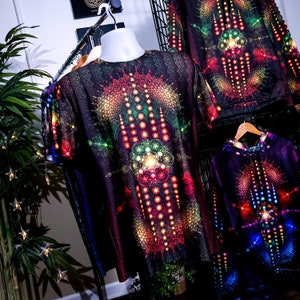 May include: Three black shirts with colorful geometric patterns. The shirts have a psychedelic, tie-dye look with a variety of colors, including red, orange, yellow, green, blue, and purple. The shirts are hanging on a rack.